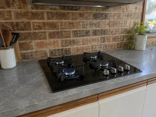 Black gas cooktop set into a gray benchtop with brick splashback and kitchen window in New Lambton Heights