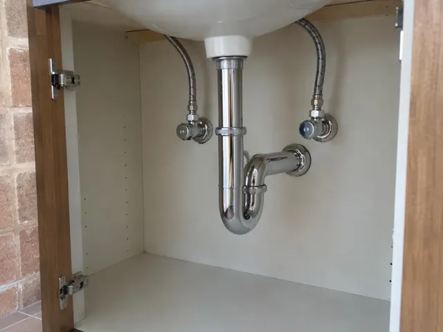 Neat under-sink plumbing with chrome trap and flex hoses inside a cabinet with white walls in Kotara